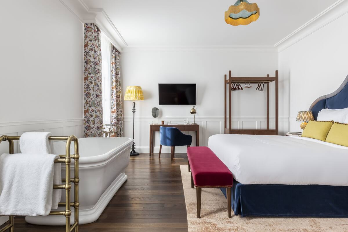 Habitación de hotel con una bañera grande, una cama con cabecera azul, un escritorio con una silla y un televisor en la pared.