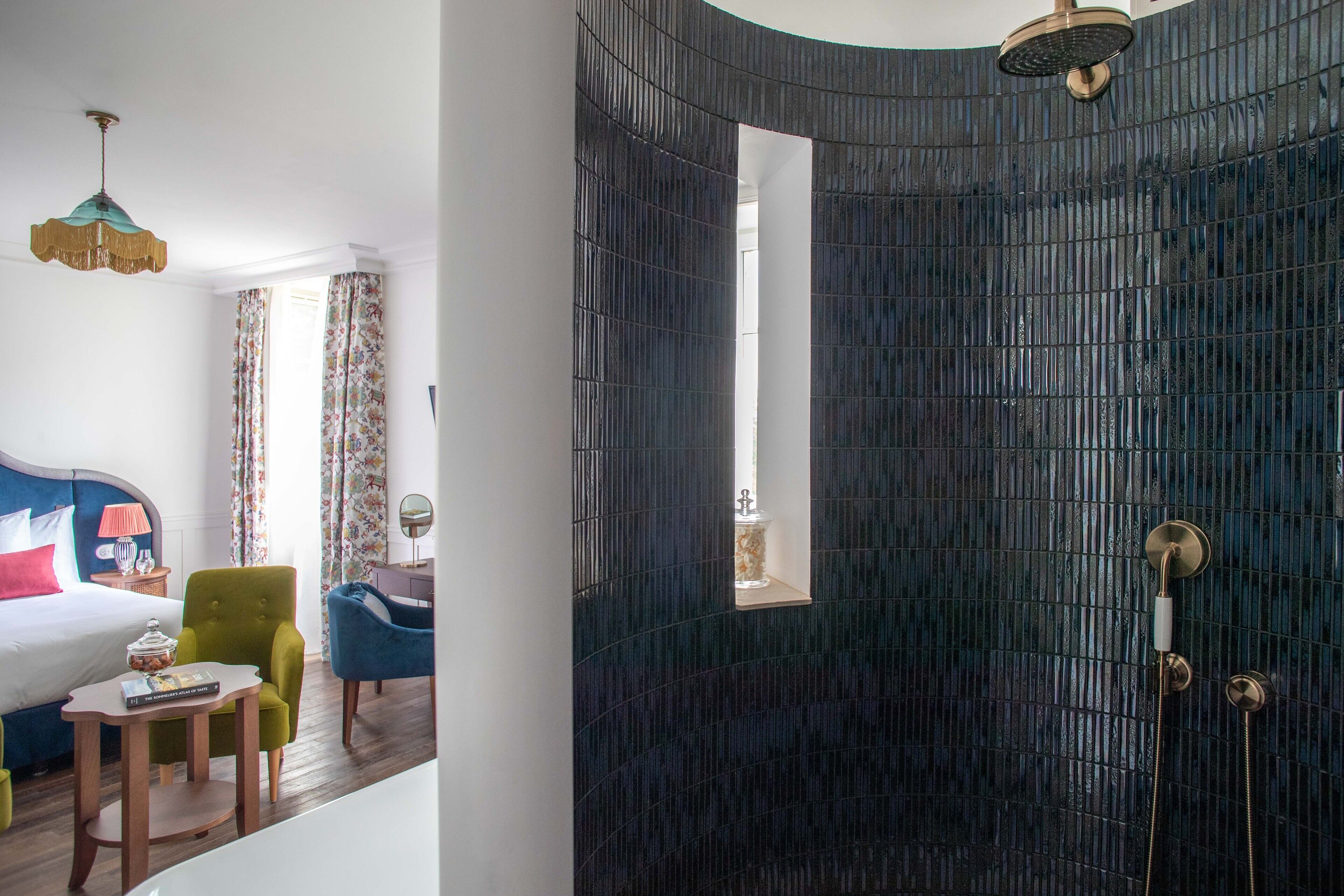 Ein Hotelzimmer mit einem Bett, einem Schreibtisch und einem Stuhl. Ein Fenster mit Vorhängen und eine Dusche mit Glastür sind ebenfalls vorhanden.