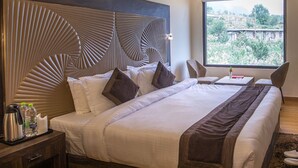 Ein Hotelzimmer mit einem großen Bett, einem Holz-Kopfteil, einem Nachttisch mit Wasserkocher und Wasserflaschen sowie einem Fenster mit Blick auf Grünflächen.