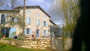 Un edificio de dos pisos con paredes de piedra y contraventanas azules, rodeado por un muro de piedra y una cerca.