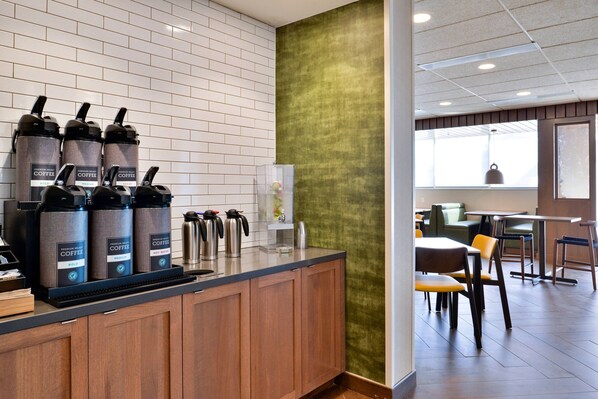 Una estación de café con varios dispensadores, un refrigerador pequeño y un área de descanso con mesas y sillas.