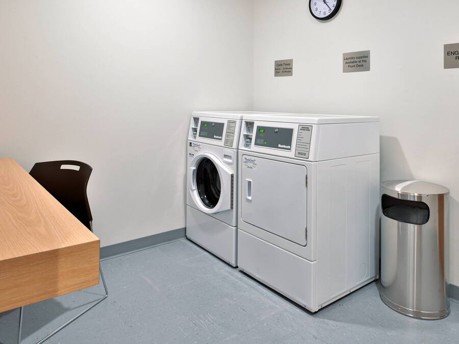 A laundry room with two front-loading washing machines, a trash can, a clock, and a table.