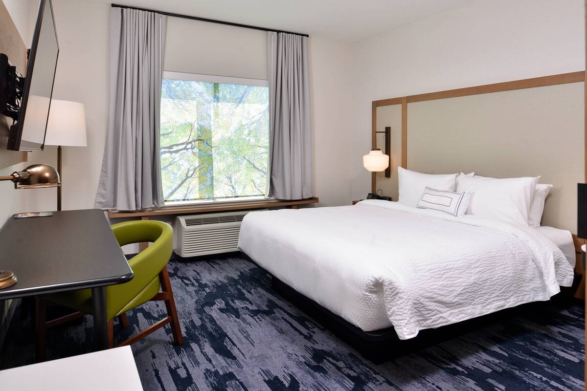 Ein Hotelzimmer mit einem großen Bett, einem Schreibtisch, einem Stuhl, einem Vorhangfenster und einer Lampe.
