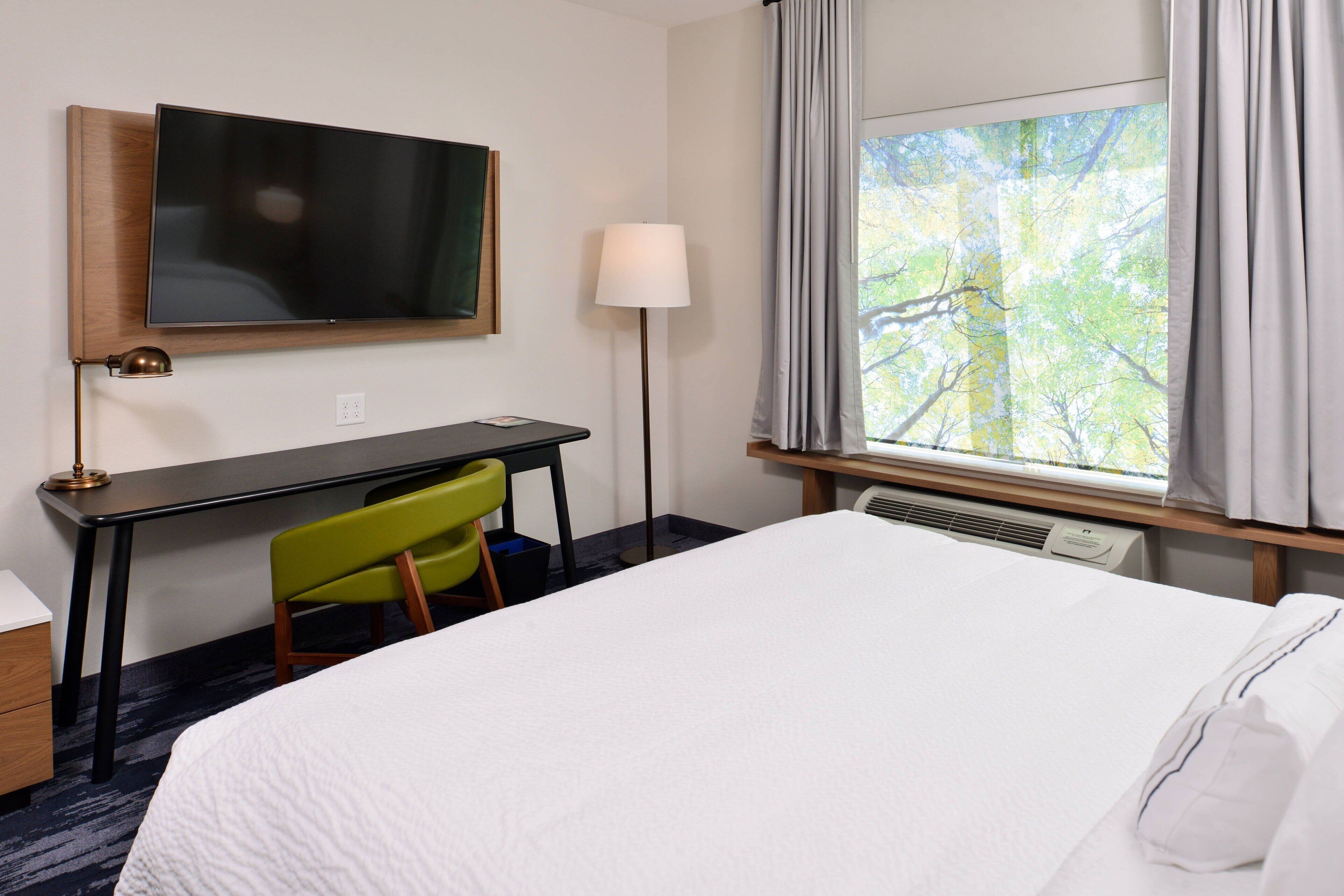 A hotel room with a bed, a desk, a chair, a television, a lamp, and a window with curtains.