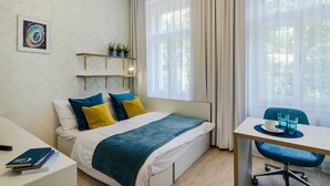 Une chambre d’hôtel moderne, équipée d’un lit, d’une table de chevet et d’un bureau avec une chaise. On y trouve une fenêtre avec des rideaux légers, une étagère fixée au mur avec des plantes et un tableau encadré.