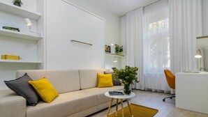 Un salon moderne comprenant un canapé, une petite table ronde, un bureau et une chaise. On y trouve des étagères ornées d’objets décoratifs et une grande fenêtre avec des rideaux.