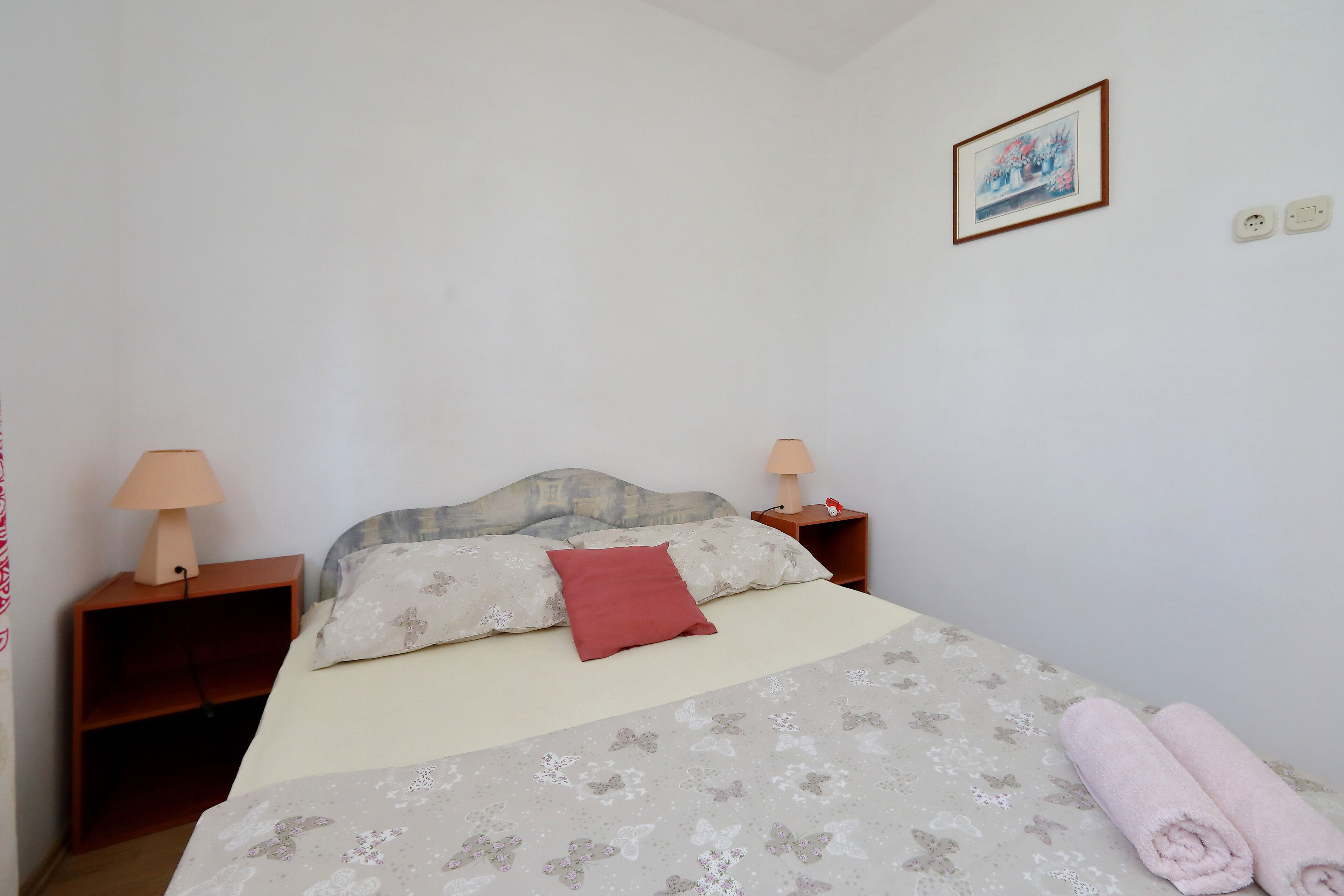A neatly made bed with a floral bedspread and a red pillow, two bedside lamps, a wooden nightstand, and a framed picture on the wall.