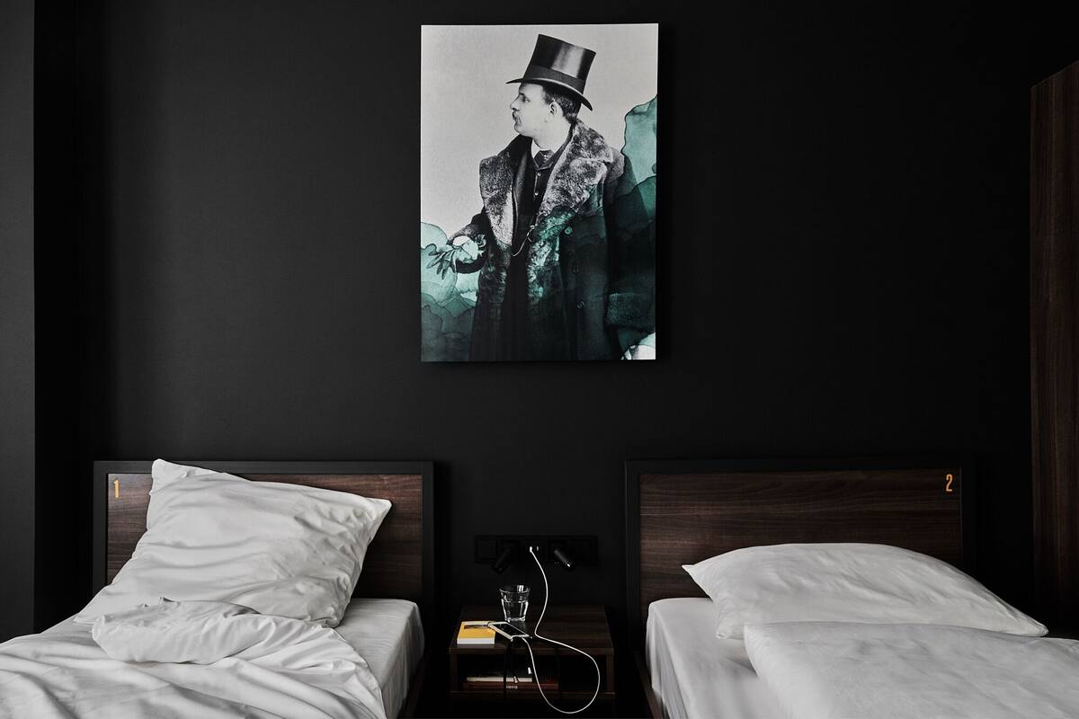 Ett hotellrum med två sängar, en mörk vägg och en inramad tavla som föreställer en person med hög hatt.