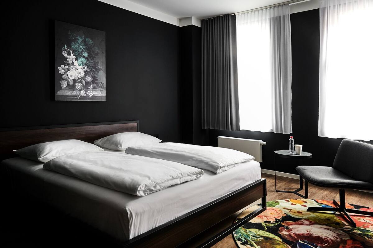Ett modernt hotellrum med en stor säng, ett litet runt bord, en stol och ett fönster med gardiner.