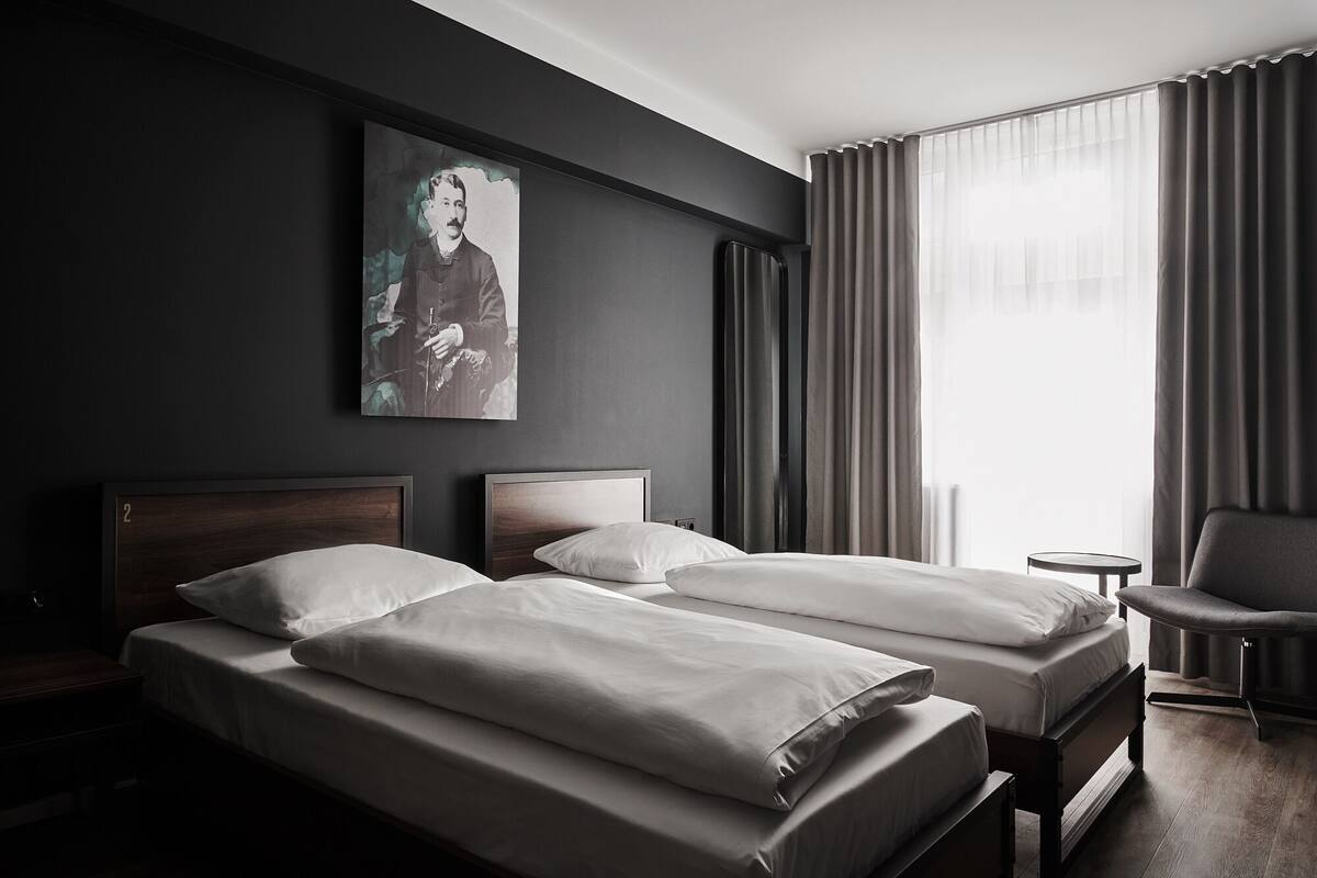 Ett hotellrum med två sängar, ett litet bord, en stol och ett stort fönster med gardiner.