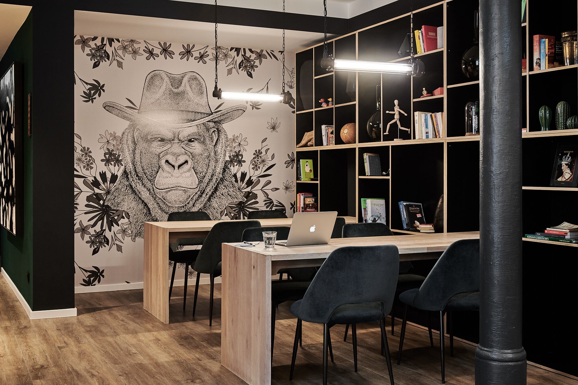 A modern office space with a wooden desk, two black chairs, a laptop, and a bookshelf filled with books and decorative items.