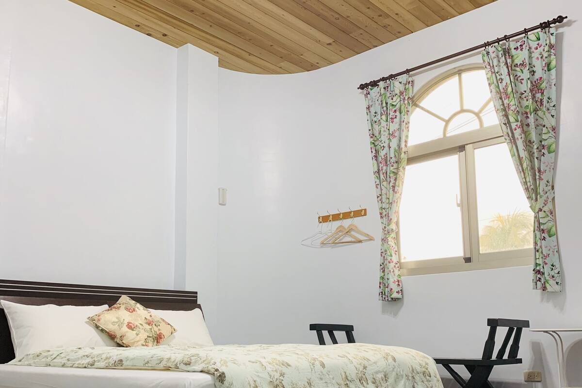 Un dormitorio con una cama, techo de madera, ventana con cortinas florales y una pequeña mesa con sillas.