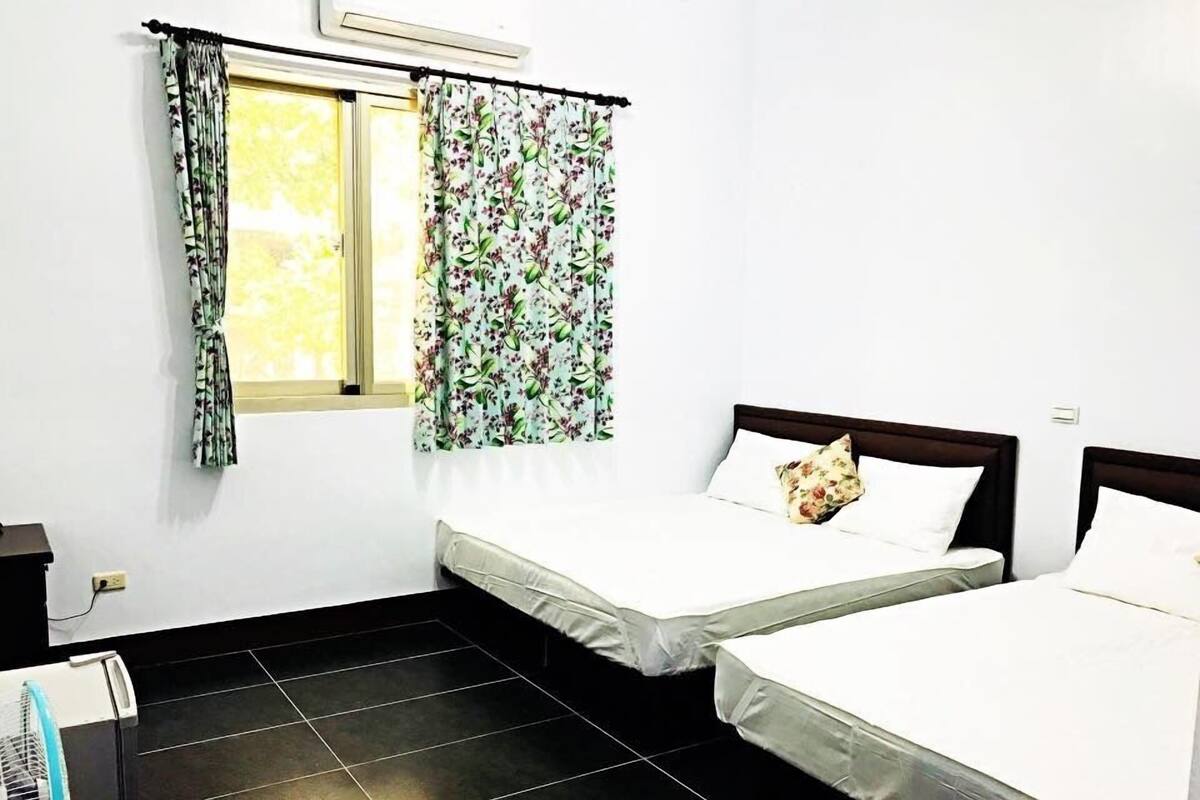 Una habitación con dos camas, una ventana con cortinas de flores, suelo de baldosas negras y un ventilador.