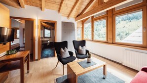 Uma sala de estar moderna com vigas de madeira, uma TV, duas poltronas, uma mesa de centro e grandes janelas com vista para uma encosta arborizada.
