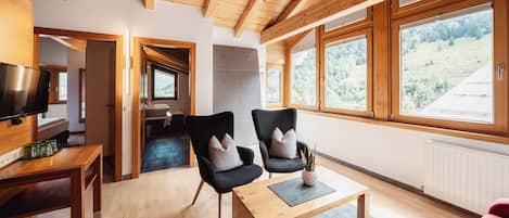 A modern living room with wooden beams, a TV, two armchairs, a coffee table, and large windows with a view of a forested hillside.