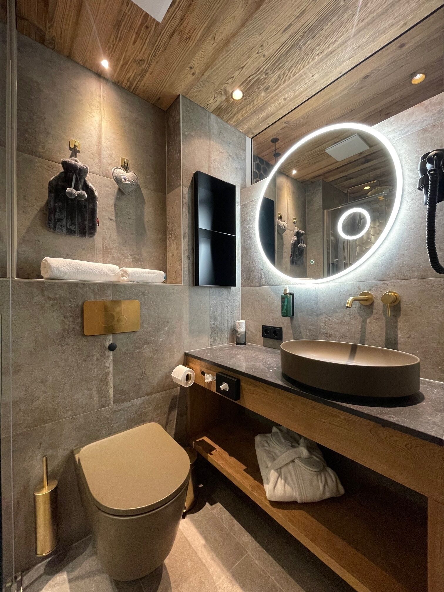 A modern bathroom with a wooden vanity, a round mirror with lights, a toilet, and a towel rack.