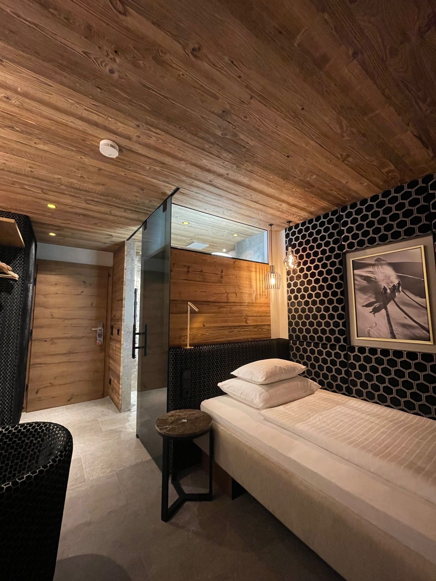 A modern bedroom with a wooden ceiling, a bed with white linens, a black and white hexagonal wall pattern, and a framed picture on the wall.