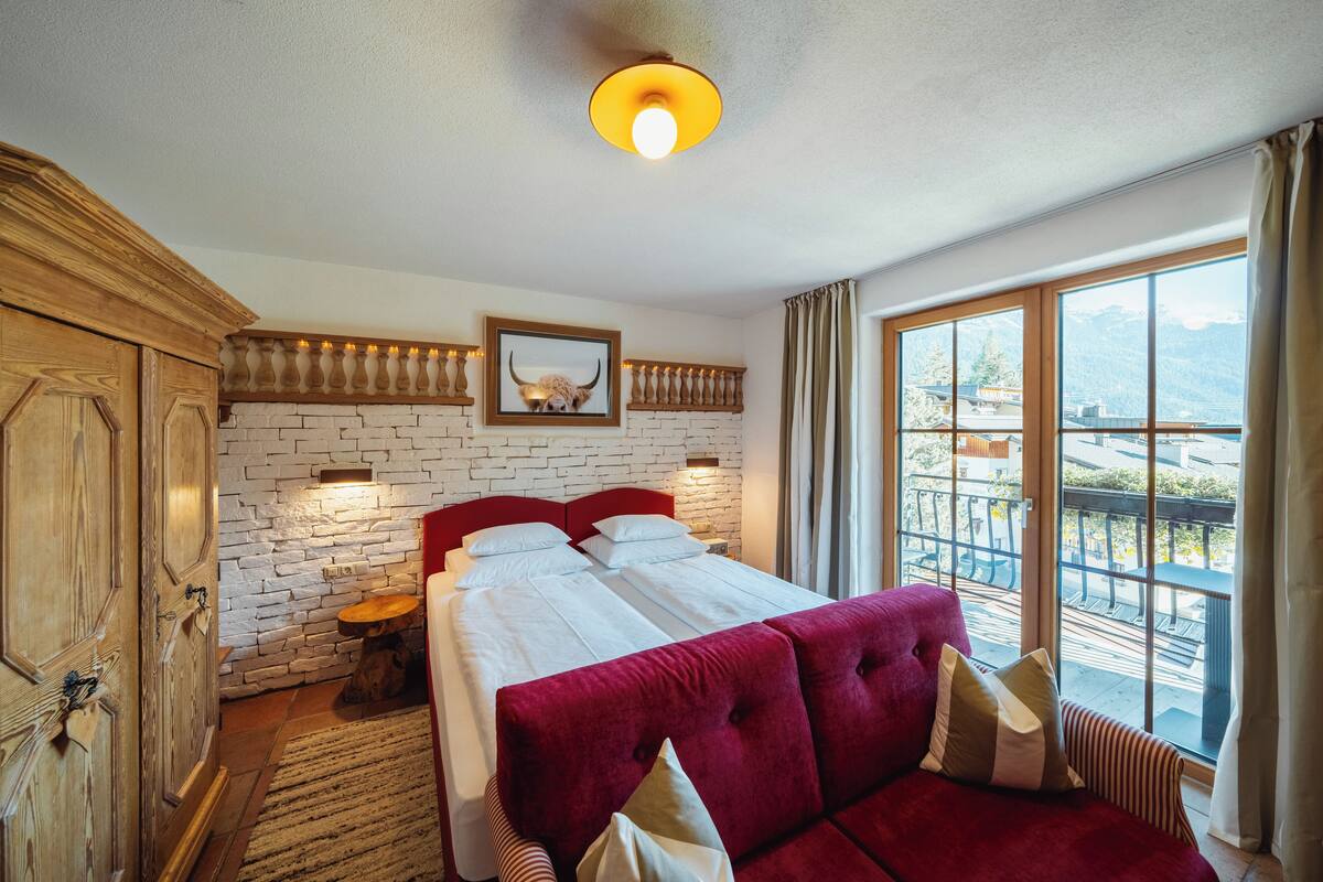 Une chambre d’hôtel avec un grand lit, une armoire en bois, un mur en briques, un tableau encadré et un balcon offrant une vue sur la montagne.