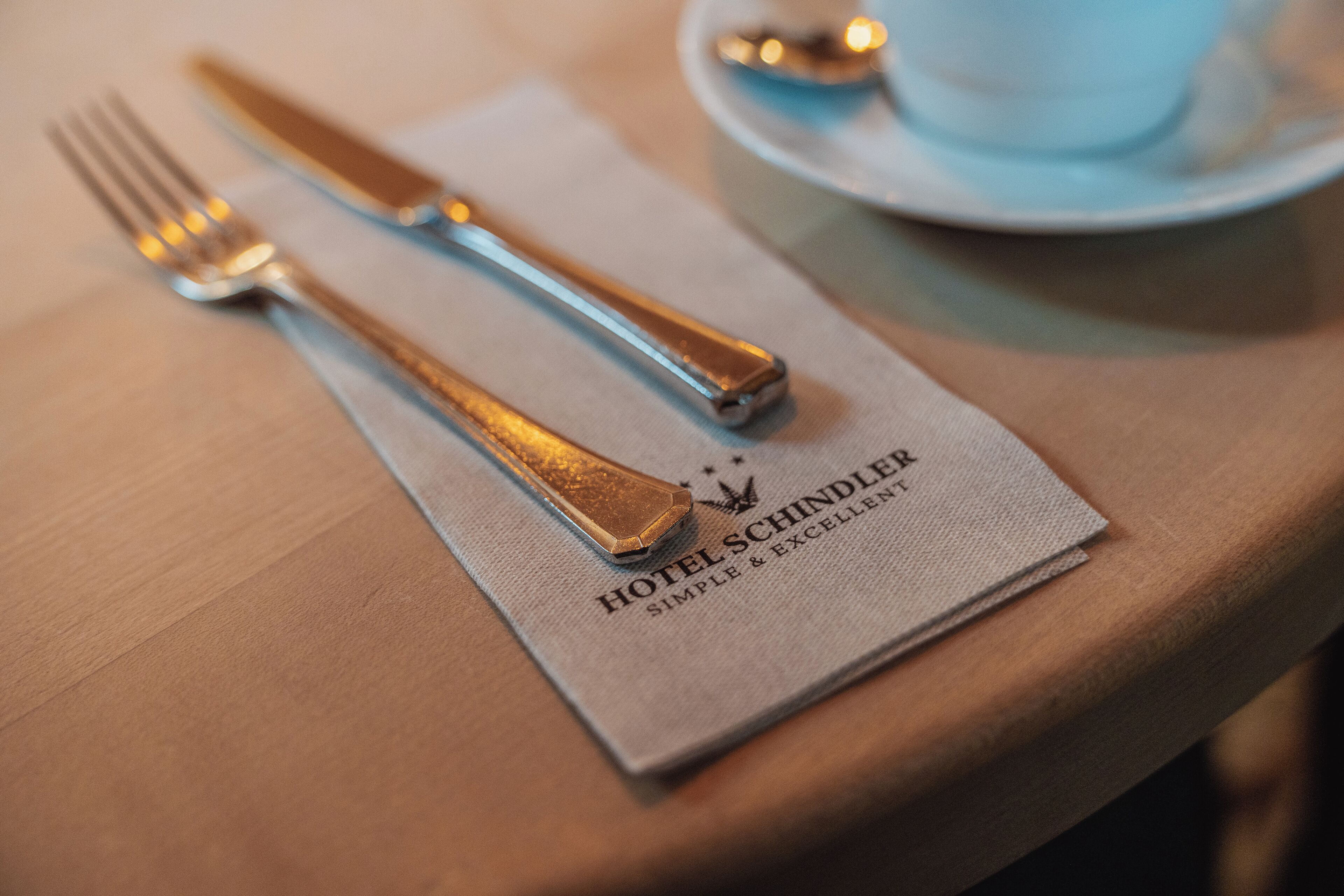 A table setting with a napkin labeled "HOTEL SCHINDLER SIMPLE & EXCELLENT" and a cup and saucer in the background.