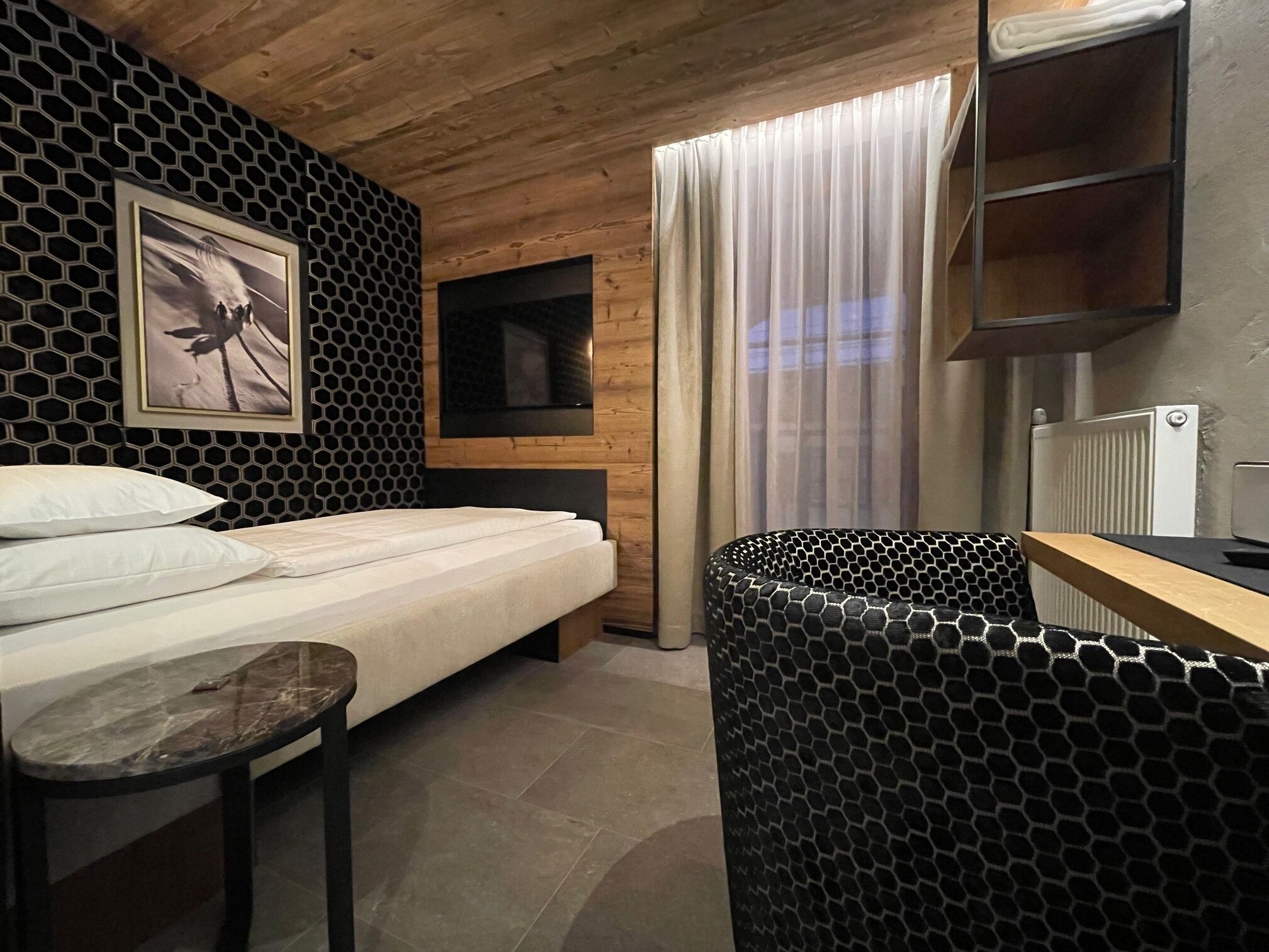 A modern bedroom with a bed, a small table, a TV, and a bathroom with a black hexagonal pattern.
