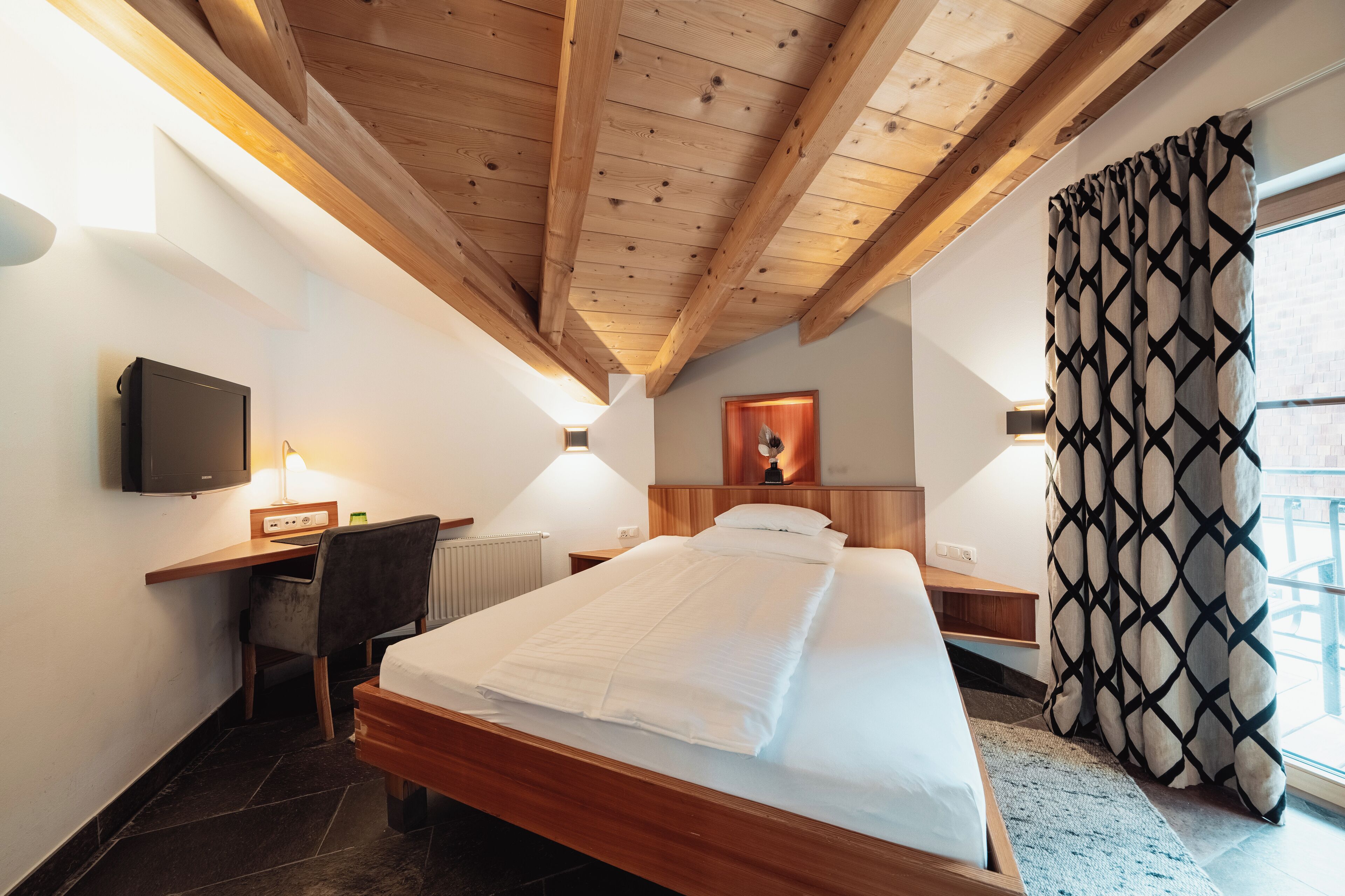 A hotel room with a wooden ceiling, a bed, a desk with a computer, a chair, a TV, and a window with curtains.