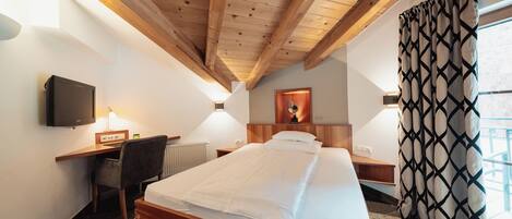 A hotel room with a wooden ceiling, a bed, a desk with a computer, a chair, a TV, and a window with curtains.