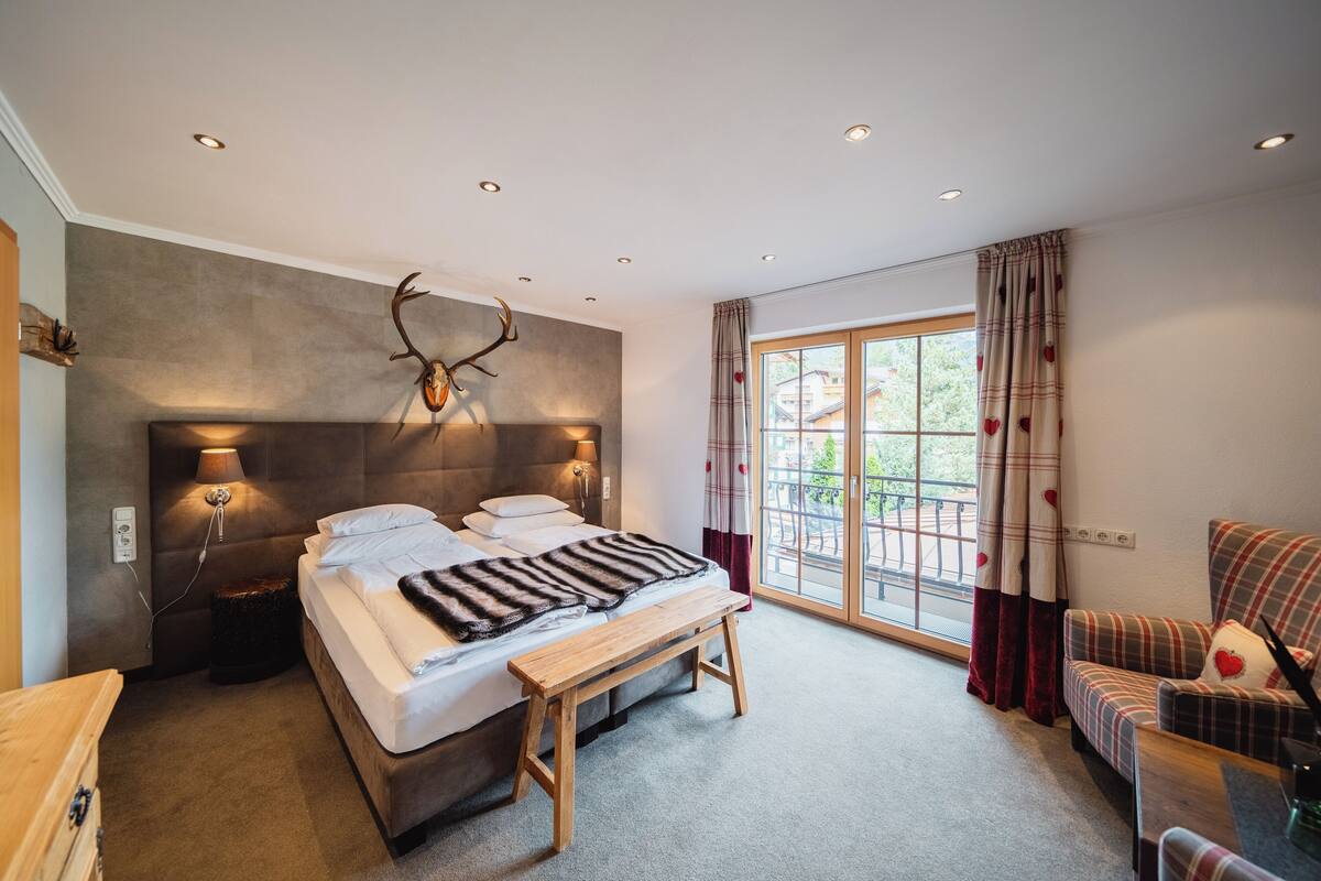 Une chambre à coucher avec un grand lit, un banc, une tête de lit ornée de bois de cerf et une vue sur l’extérieur par une fenêtre.