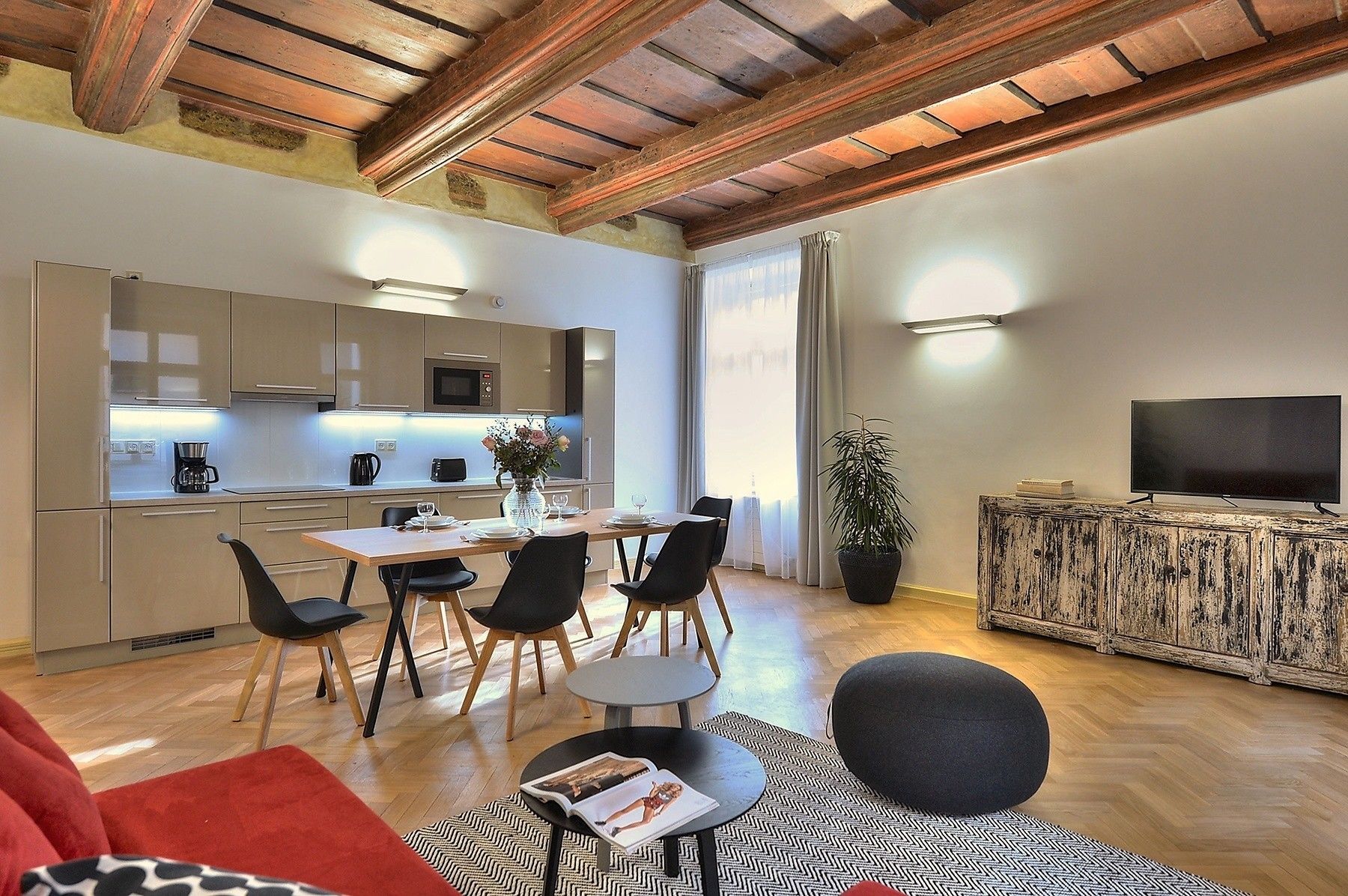 Un salon moderne avec un plafond en bois, un coin repas comprenant une table et des chaises, un téléviseur posé sur un meuble en bois et une grande plante en pot.