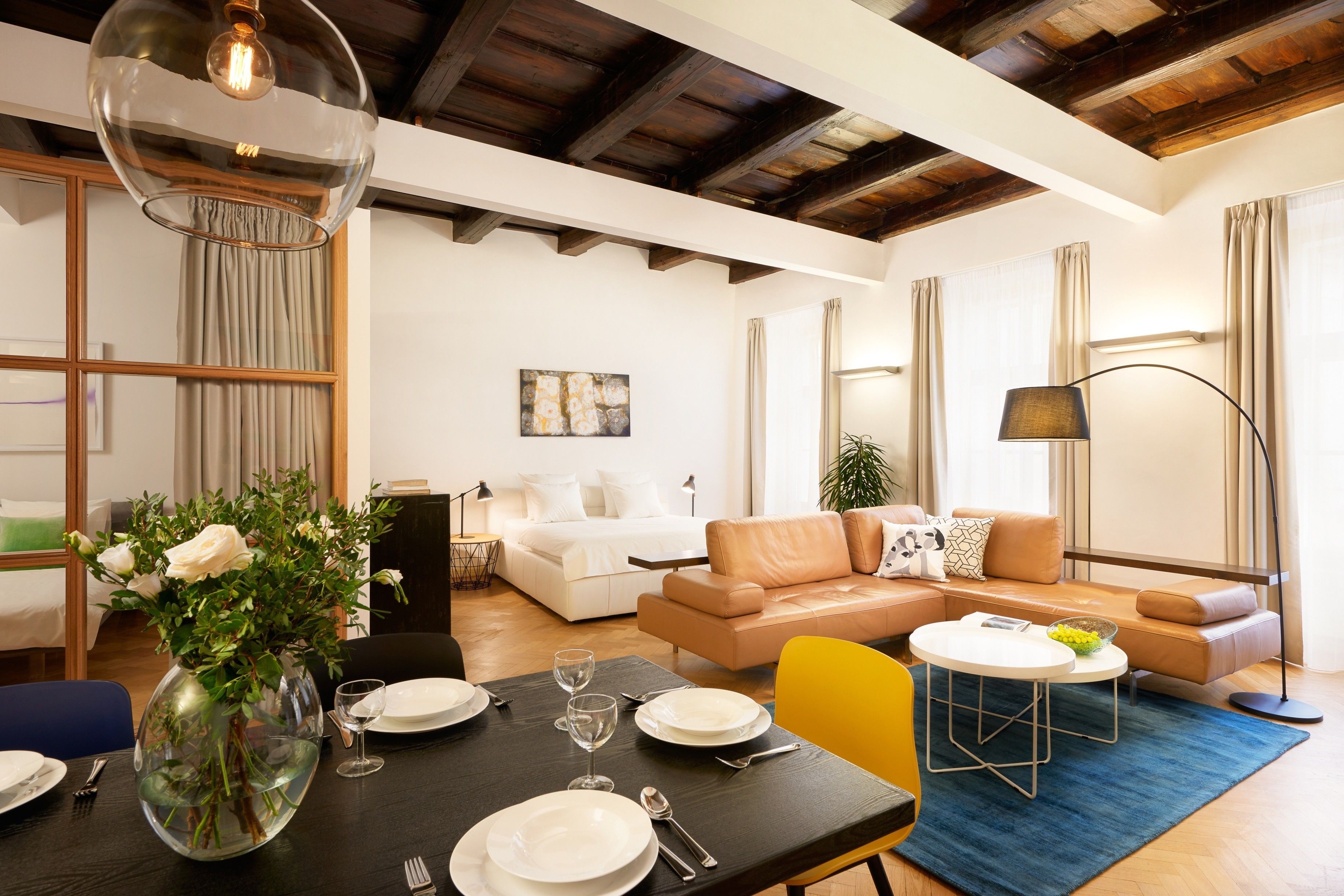 A modern living room with a dining area, featuring a wooden ceiling, a sofa set, a coffee table, and a vase with flowers.
