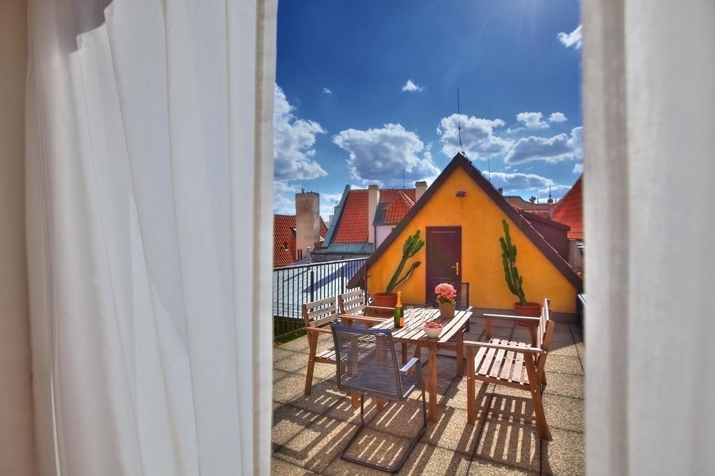 Ein Balkon mit Tisch und Stühlen, eine gelbe Wand mit einem Kaktusgemälde und im Hintergrund Gebäude mit roten Dächern.
