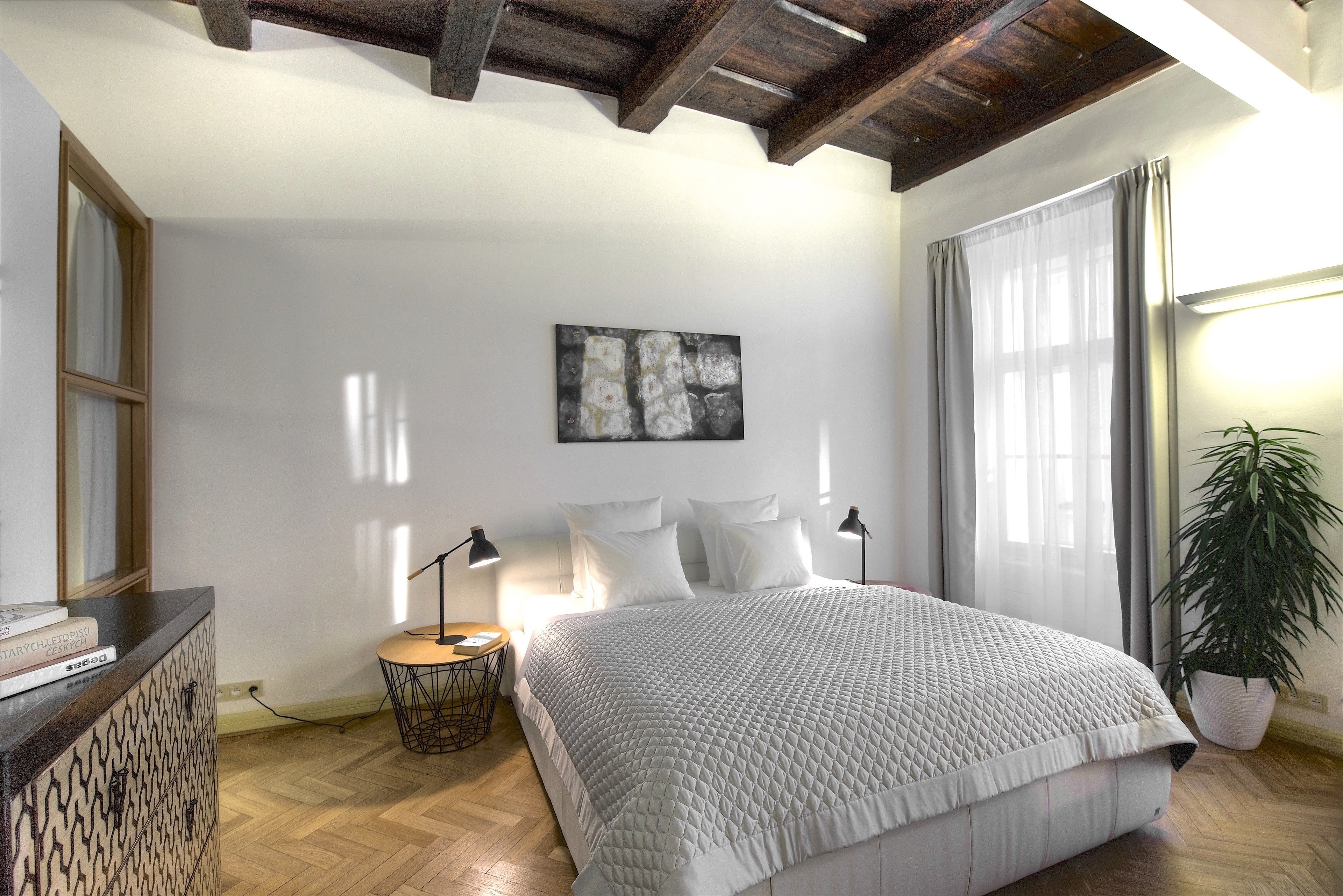 A bedroom with a large bed, a bedside table, a lamp, a plant, and a painting on the wall.