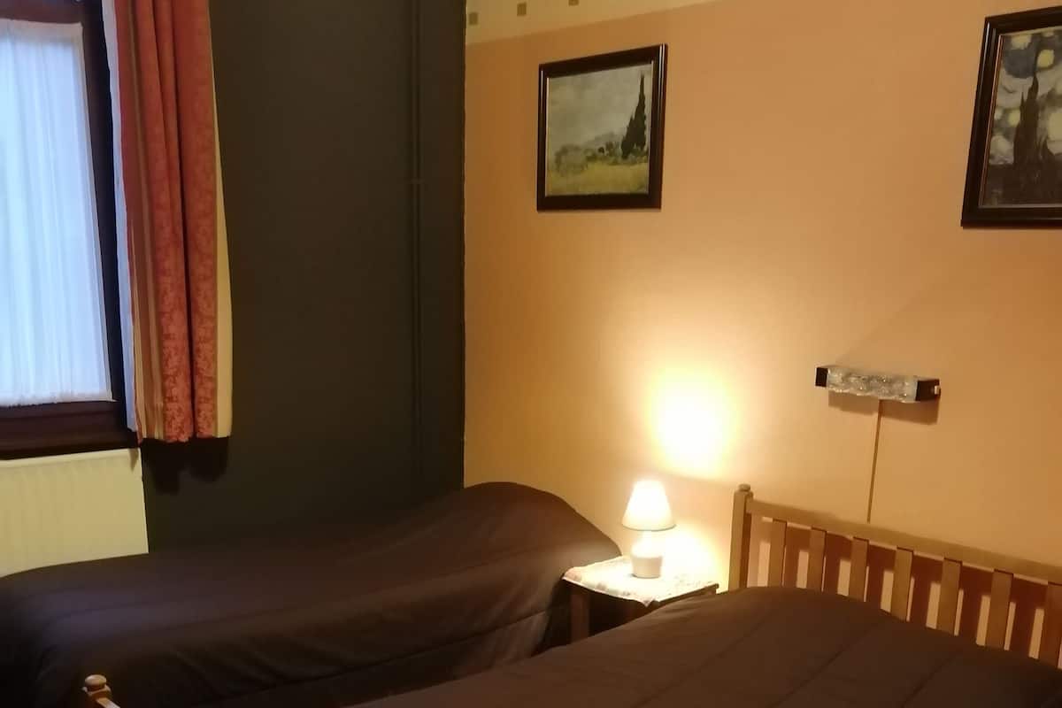 A hotel room with two single beds, a nightstand with a lamp, a window with curtains, and a wall-mounted picture.