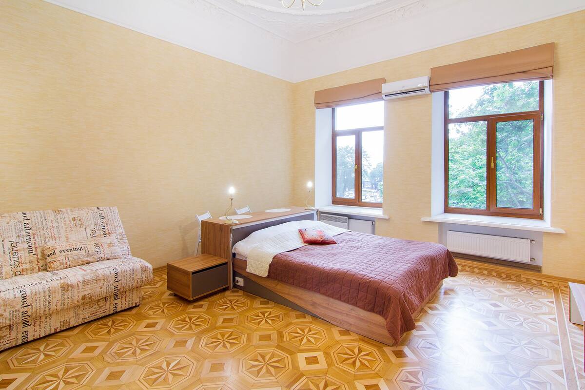 A bedroom with a bed, a sofa, a window with a view of trees, and a patterned floor.