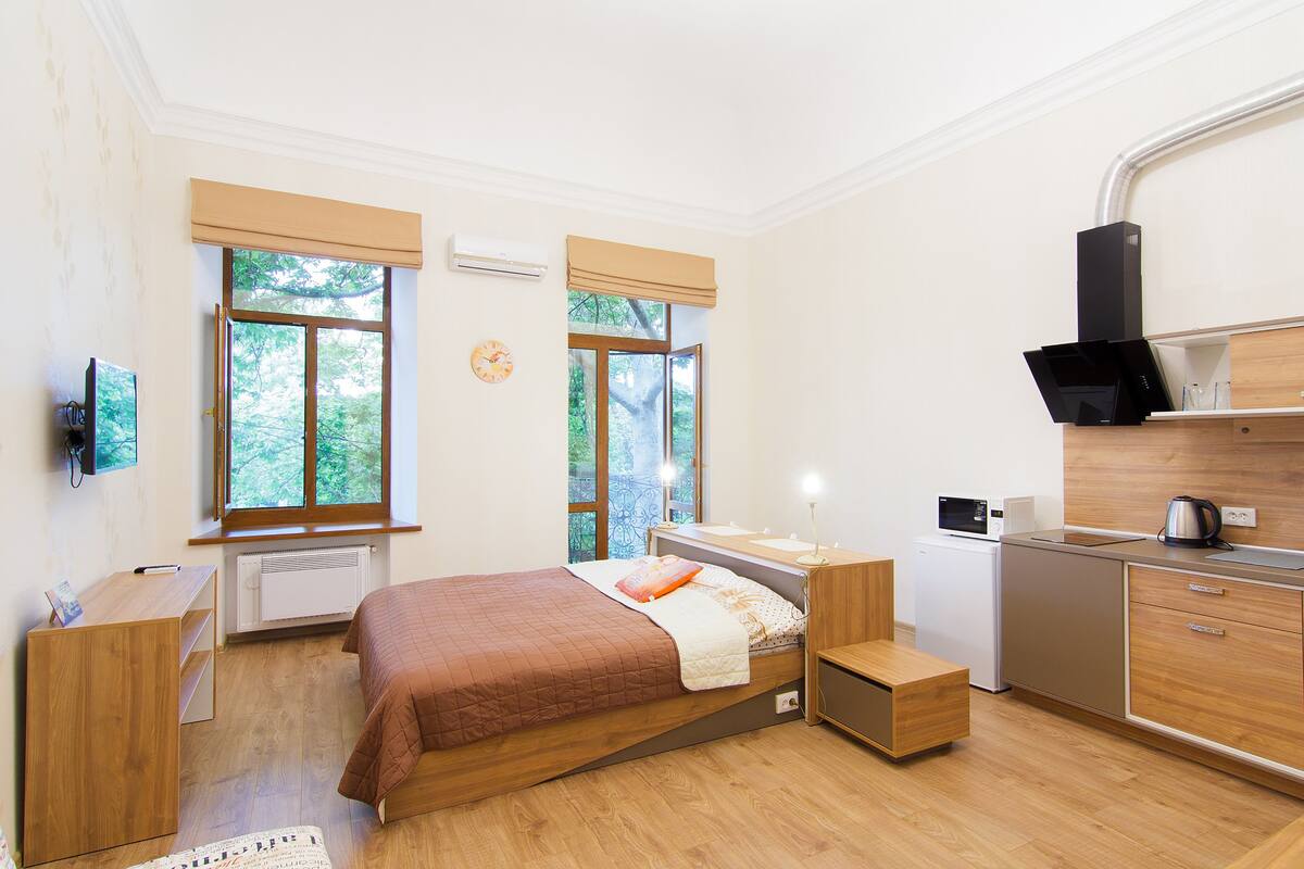 A bedroom with a bed, bedside table, and a small desk. There is a window with a view of trees, a radiator, and a clock on the wall.
