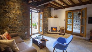 A cozy living room with a stone wall, wooden beams, a fireplace, and a blue chair. - Lyhnos (Askas)