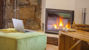 A cozy living room with a stone wall, a fireplace with a visible fire, a laptop on a green ottoman, and a wooden table with a plate of cookies. - Lyhnos (Askas)