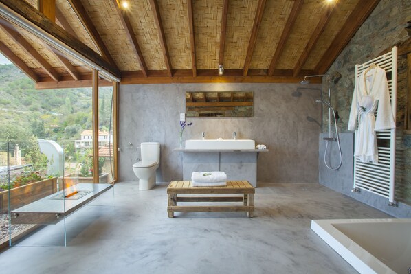 A modern bathroom with a wooden bench, a large window, a stone wall, and a shower area.