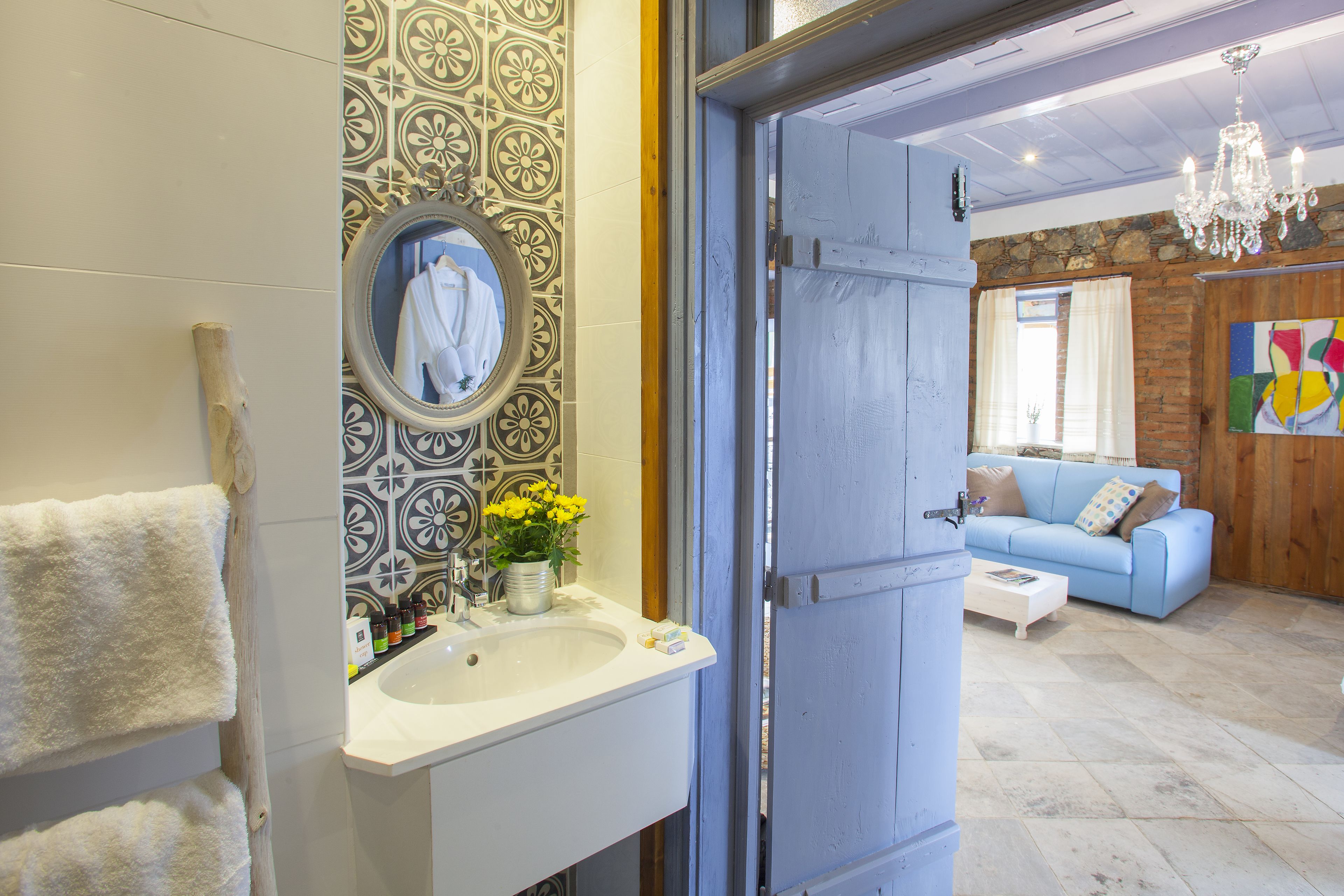 A bathroom with a sink, mirror, and towel rack. There is a door leading to a living area with a sofa and a chandelier.
