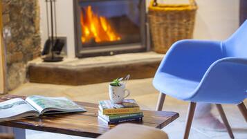 A cozy living room with a fireplace, a blue chair, a coffee table with books and a cup, and a stone wall.
