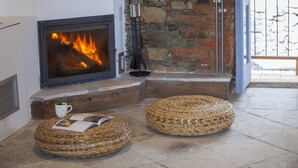A fireplace with a fire, two woven ottomans, a white mug, and a book on the floor. - Lyhnos (Askas)