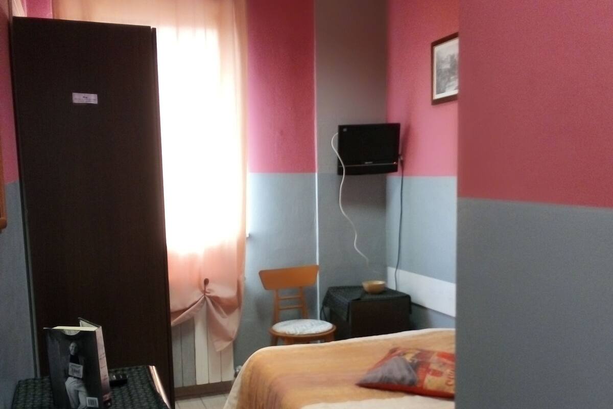 A hotel room with a bed, a desk, a chair, a television mounted on the wall, and a window with curtains.