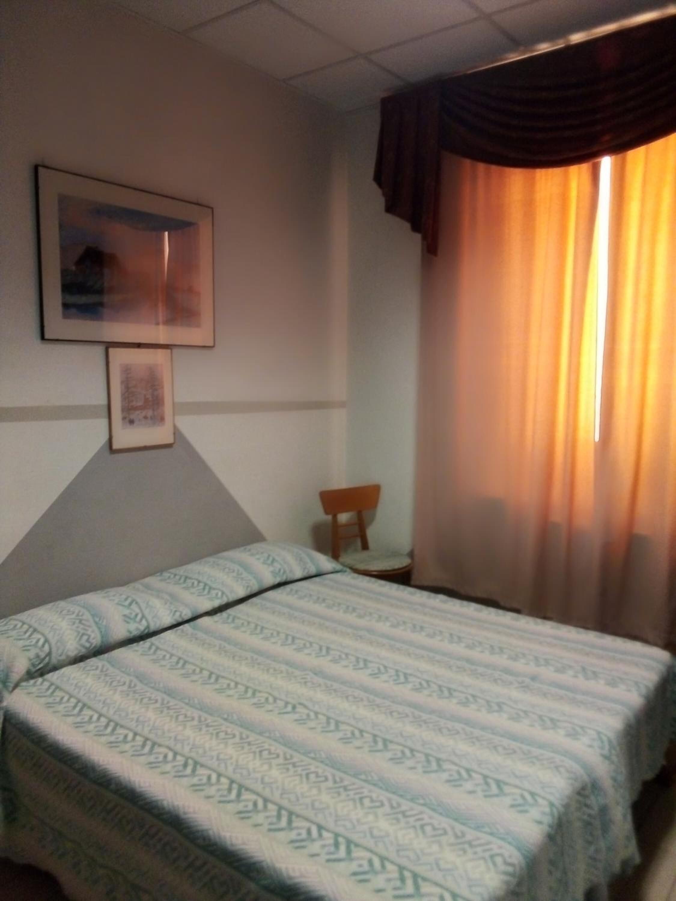 A hotel room with a bed, a chair, a painting, and a window with curtains.