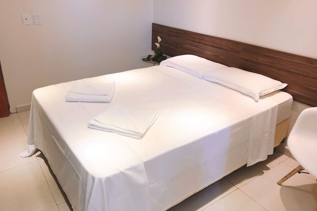 A neatly made bed with white linens, a wooden headboard, and a white chair beside it.