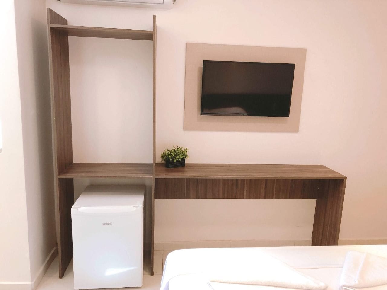 Quarto com cama, escrivaninha, pequeno refrigerador e TV fixada na parede.