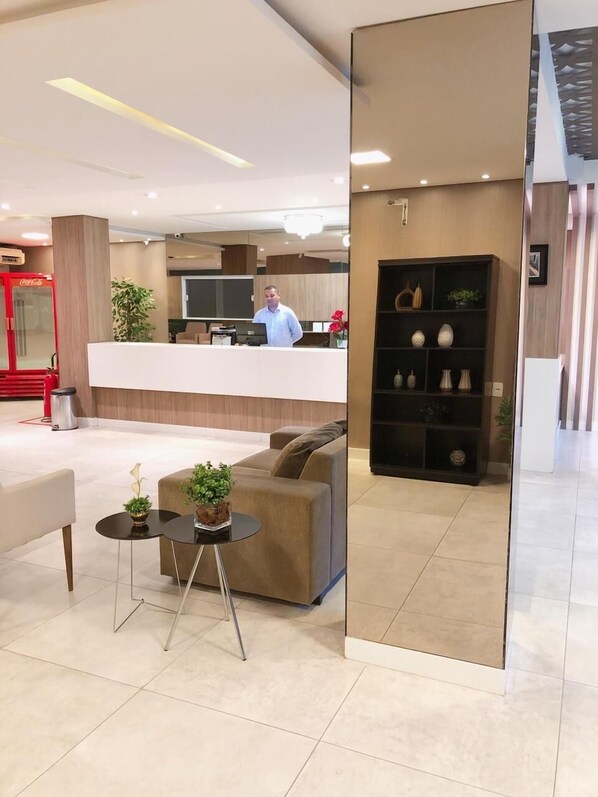 A modern hotel lobby with a reception desk, a seating area with a sofa and two small tables, and a decorative bookshelf.