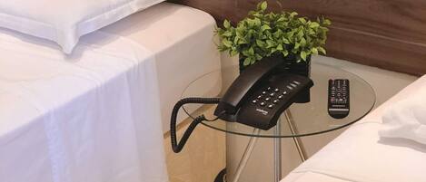 Habitació d'hotel amb un llit, una taula de nit amb un telèfon i comandament a distància, i una petita planta en test.