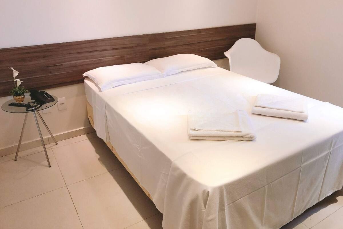 A neatly made bed with white linens, a small table with a phone and a plant, and a white chair in a simple room with a wooden headboard.