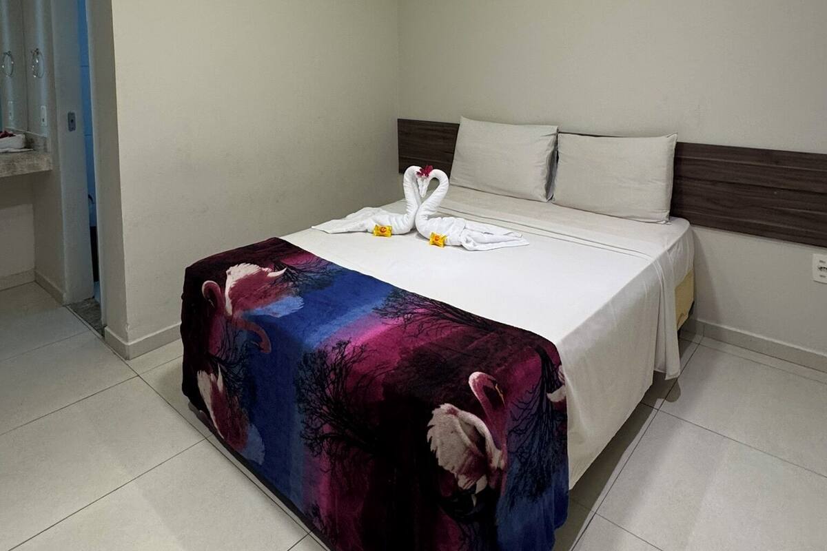 A neatly made bed with a white comforter and two swan-shaped towels. The room has a modern design with a dark headboard and a colorful blanket on the bed.