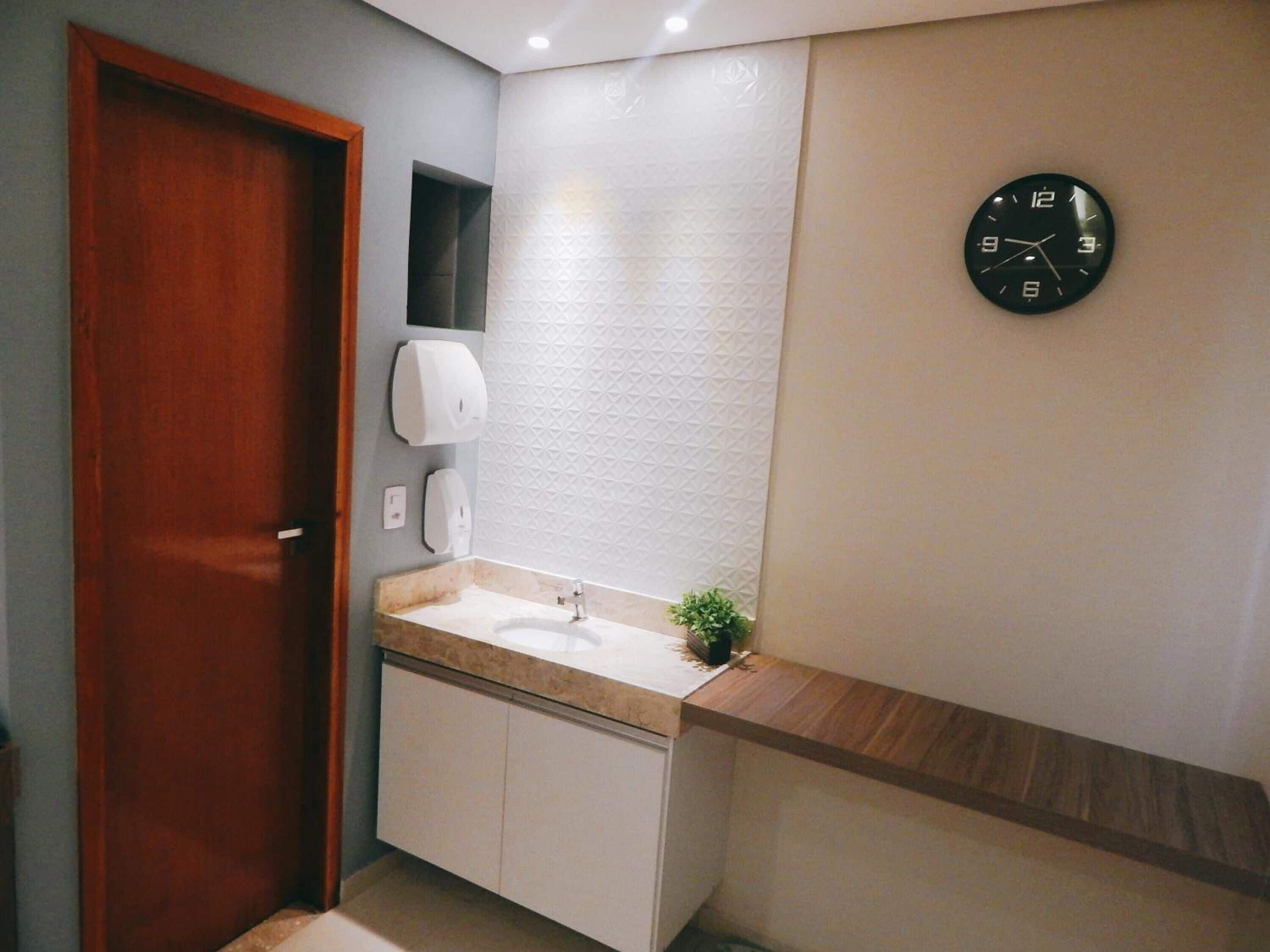 A bathroom with a wooden door, a sink, a mirror, a clock, and a soap dispenser.