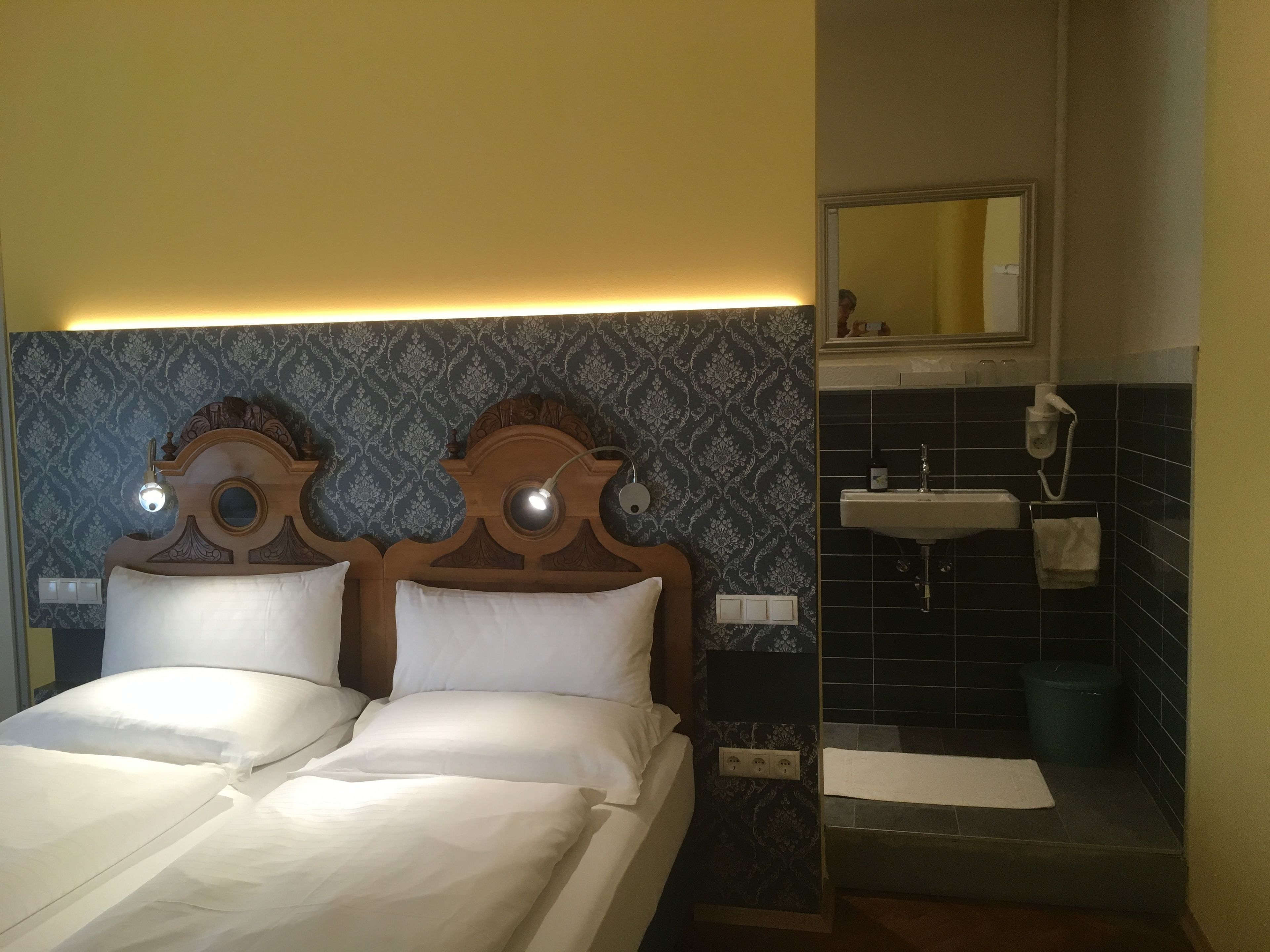 Hotelkamer met een tweepersoonsbed, een houten hoofdbord, nachtkastje, wastafel en een prullenbak.